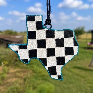 May include: Texas-shaped car air freshener with a black and white checkered pattern. The outline is trimmed with a sparkling blue material, and it hangs from a black cord. The background shows a blurred outdoor scene.