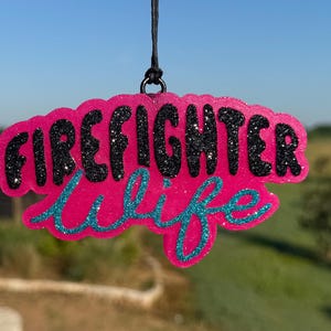 May include: A hanging ornament with a bright pink glitter base. The word FIREFIGHTER is written in bold, black glittery capital letters, and the word wife is written below in a cursive, teal glittery script. It hangs from a thin black cord.