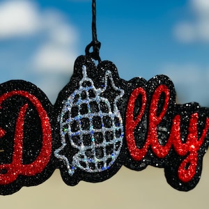 May include: A black glittery ornament with the name "Dolly" in red glitter script. A silver disco ball design is in the center. The ornament hangs from a black string against a blue sky background.