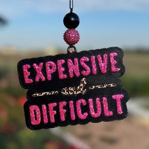 May include: Black and pink glittery car air freshener with the words "EXPENSIVE" and "DIFFICULT". The air freshener is suspended by a black string and decorative beads.