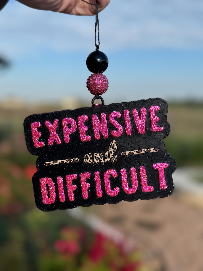 May include: Black and pink glittery car air freshener with the words "EXPENSIVE" and "DIFFICULT". The air freshener is suspended by a black string and decorative beads.