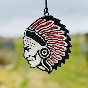 May include: A decorative air freshener featuring a Native American chief design. The image has a black outline with red and white accents. The air freshener is hanging from a black string against a blurred green and gray background.