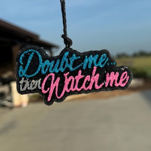 May include: A black car air freshener with the text "Doubt me... then watch me" in pink and blue glitter.