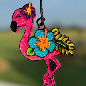 May include: Pink flamingo ornament with glitter, decorated with blue, green, orange and purple flowers. The ornament is hanging from a black string.