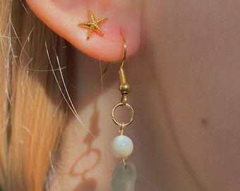 May include: Gold earrings with a white shell and a light blue stone dangle from a gold hook. A small gold starfish stud earring is in the other ear.