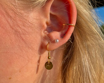 May include: A close-up of a person's ear with gold hoop earrings and a gold dangle earring with a small round charm. The dangle earring has a small gold bead at the top.