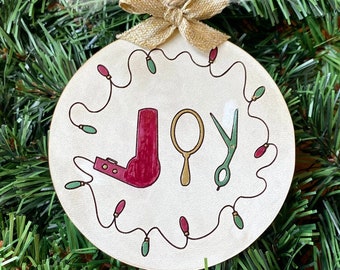 May include: A round, cream-colored Christmas ornament with the word "JOY" and illustrations of a hairdryer, mirror, and scissors. The ornament is decorated with a string of lights and a gold bow. The text "SVG FILE" is at the top.