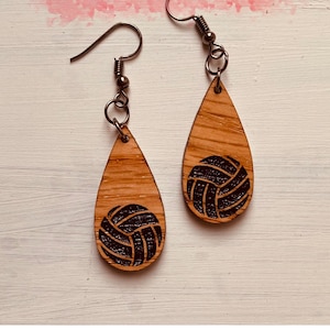 Puede incluir: Un par de pendientes de madera en forma de lágrima con un diseño de voleibol negro. Los pendientes están sobre un fondo de madera blanco.