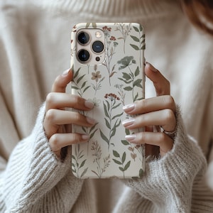 May include: A phone case with a floral design. The case features a cream-colored background with a pattern of green, brown, and red botanical illustrations. The phone case is held by a person wearing a cream-colored sweater.