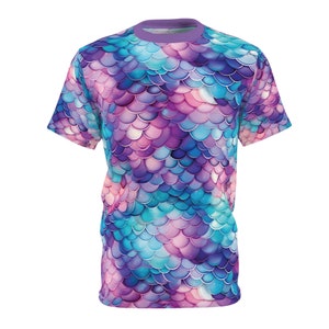 Puede incluir: Una camiseta de manga corta con un estampado vibrante en toda la superficie. El diseño presenta un patrón de escamas en tonos de azul, morado y rosa, creando una apariencia texturizada. La camiseta tiene cuello redondo con ribete morado.