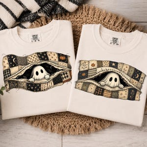 Puede incluir: Dos camisetas de cuello redondo en color blanco roto con un gráfico de un fantasma asomándose desde debajo de una colcha de retazos. El diseño de la colcha incluye varios patrones como estrellas, lunas y calabazas en tonos negro, tostado y crema. Las camisetas tienen una etiqueta Comfort Colors en el cuello.