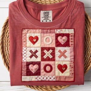 May include: A red t-shirt with a patchwork design featuring hearts and X's and O's. The shirt is folded and placed in a woven basket. The shirt has a Comfort Colors tag.