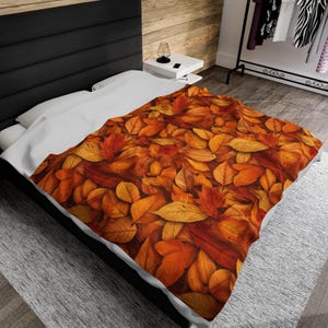 May include: A bed with a white duvet cover and a brown and orange patterned throw blanket. The blanket features a close-up image of autumn leaves.