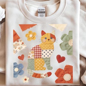 May include: Cream-colored sweatshirt featuring a patchwork cat design with floral and polka dot patterns. The cat is surrounded by fabric flowers and hearts in various colors, creating a whimsical, handcrafted aesthetic.