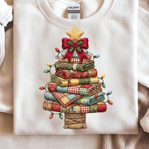 May include: Cream-colored sweatshirt featuring a Christmas tree design. The tree is constructed from stacked, patterned quilts, adorned with a red bow, star, and string lights. The base of the tree is a burlap sack.