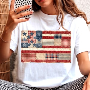 Puede incluir: Una camiseta blanca de manga corta con un gráfico rectangular de una bandera estadounidense en estilo patchwork. El diseño de la bandera se compone de varios patrones de tela, incluyendo cuadros vichy rojos y blancos, mezclilla azul y rayas.