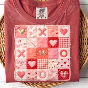 May include: A red-toned t-shirt with a patchwork design featuring hearts, X's, and O's in various shades of pink and red. The design includes floral and gingham patterns, with gold glitter accents. The shirt has a Comfort Colors label.
