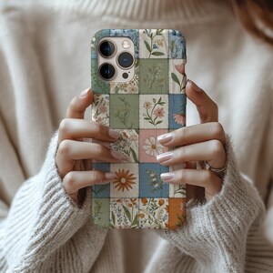 May include: A phone case with a patchwork design showcasing floral patterns in a variety of colours. The case features square panels with botanical illustrations in shades of pink, blue, green, and orange. The case is held by a person wearing a cream-coloured sweater.