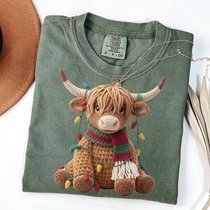 May include: Sage green t-shirt with a crocheted Highland cow design, wearing a festive scarf and fairy lights. The cow has brown hair, white horns, and a red and green scarf. A silver necklace is draped across the shirt. The t-shirt is a Comfort Colors brand.