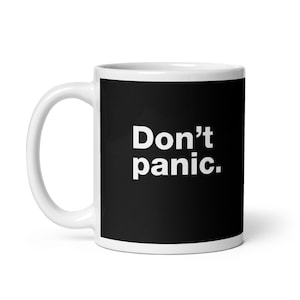 May include: A white ceramic mug featuring a black rectangular panel on the front. The text Don't panic. is printed in bold white sans-serif letters centred within the black area. The mug has a curved handle on the left side.