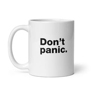 May include: A white ceramic mug with a handle on the left side. The text Don't panic. is printed in bold black sans-serif font on the front of the mug. The mug has a smooth, glossy finish and a cylindrical shape with a flat base.
