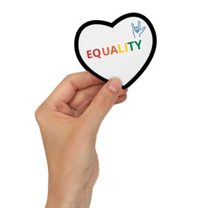 Peut inclure: Une carte en forme de cœur blanche avec une bordure noire. La carte porte le mot "ÉGALITÉ" écrit en couleurs arc-en-ciel et un signe de la main bleu pour "Je t'aime" en langue des signes française.