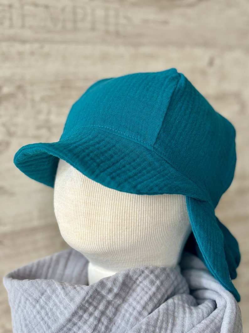 Puede incluir: Un sombrero de pescador color verde azulado hecho de una tela texturizada y arrugada. El sombrero presenta un ala ancha inclinada hacia abajo y una copa redondeada. Se muestra sobre una cabeza de maniqu&iacute; color hueso, con una tela gris suave drapeada en la base.