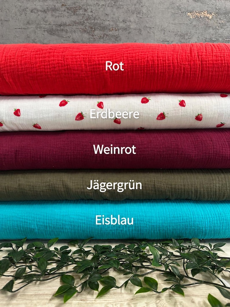 Puede incluir: Cinco rollos apilados de tela texturizada en rojo s&oacute;lido, blanco con un estampado de fresas peque&ntilde;as, rojo vino, verde oliva y azul cian brillante. El texto Rot, Erdbeere, Weinrot, J&auml;gergr&uuml;n y Eisblau est&aacute; impreso sobre cada rollo de tela.