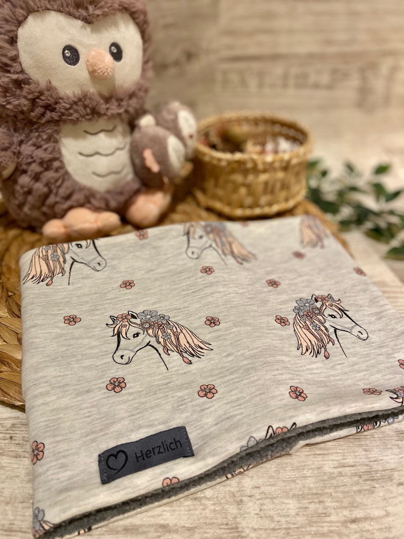 May include: A grey and white fabric neck warmer with a floral pattern featuring horses with flower crowns. The neck warmer has a grey fleece lining and a black label with a heart and the word "Herzlich".