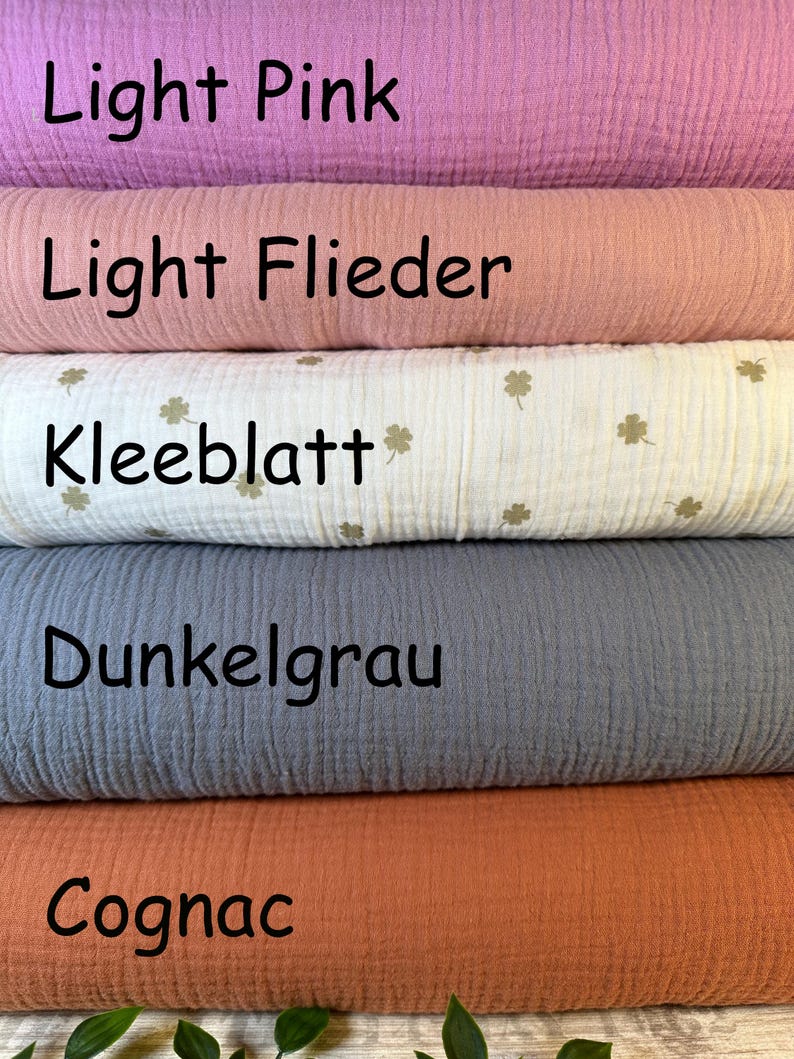 Puede incluir: Cinco rollos apilados de tela de muselina texturizada en varios colores. El rollo superior es rosa claro, seguido de lila claro, un rollo blanco con un peque&ntilde;o patr&oacute;n de tr&eacute;bol dorado etiquetado como Kleeblatt, un rollo gris oscuro etiquetado como Dunkelgrau y un rollo color co&ntilde;ac.