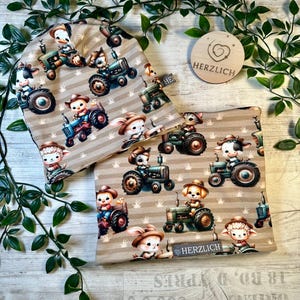 Könnte beinhalten: Ein beiges Mützen- und Halstuch-Set mit einem sich wiederholenden Muster von Zeichentricktieren, die Traktor fahren. Zu den Tieren gehören eine Kuh, ein Hase, ein Hund und ein Schaf. Der Text "HERZLICH" ist sichtbar.