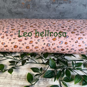 Puede incluir: Un rollo de tela con fondo rosa claro y un patr&oacute;n de leopardo repetitivo en tonos naranja, gris y negro. El texto "Leo hellrosa" est&aacute; impreso en letras verde oscuro a trav&eacute;s del centro del rollo de tela.