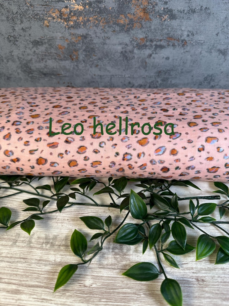 Puede incluir: Un rollo de tela con fondo rosa claro y un patr&oacute;n de leopardo repetitivo en tonos naranja, gris y negro. El texto "Leo hellrosa" est&aacute; impreso en letras verde oscuro a trav&eacute;s del centro del rollo de tela.