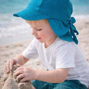 Könnte beinhalten: Ein Kind mit einem türkisfarbenen Sonnenhut mit Krempe und Bindeband im Nacken spielt im Sand. Das Kind trägt ein weißes T-Shirt und türkisfarbene Shorts. Der Hintergrund zeigt das Meer und den Himmel.
