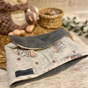 May include: A white and grey reversible neck warmer with a unicorn print and pink flowers. The warmer has a grey fleece lining and a label that says "Herzlich".