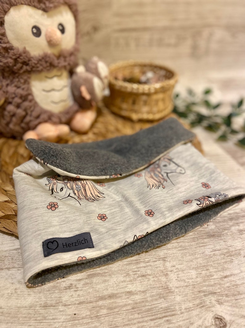 May include: A white and grey reversible neck warmer with a unicorn print and pink flowers. The warmer has a grey fleece lining and a label that says "Herzlich".
