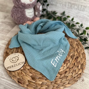 Puede incluir: Un babero de muselina azul claro con el nombre "Emely" bordado en blanco. El babero está sobre una estera de paja tejida con un letrero de madera que dice "HERZLICH" con un corazón.