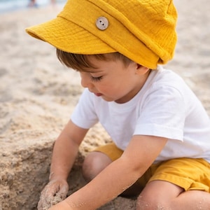 Könnte beinhalten: Eine senfgelbe Mütze im Stil eines Zeitungsjungen mit einem Holzknopf. Der Hut ist aus einem strukturierten Stoff gefertigt. Die Person trägt ein weißes T-Shirt und gelbe Shorts. Die Person spielt im Sand am Strand.