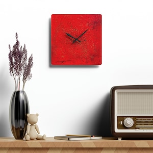 May include: A red square clock with black hands on a white wall. The clock is surrounded by a white wall, a vase with purple flowers, a wooden teddy bear, a book, and a pencil. There is also a vintage radio on the shelf.