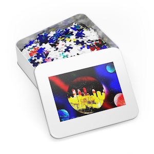 May include: A white metal tin with a colorful cityscape puzzle inside. The puzzle depicts a city skyline inside a red and yellow sphere against a dark blue background with white stars and three planets.