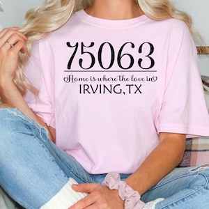 May include: A light pink t-shirt with the number "75063" printed in large black letters. Below the number is the phrase "Home is where the love in" and "IRVING, TX" in black script. The shirt is a casual style.