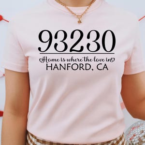 May include: A light pink t-shirt with the number "93230" printed in black, along with the text "Home is where the love in HANFORD, CA". The shirt is short-sleeved.