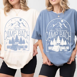 May include: Two women wearing oversized t-shirts with a camping theme graphic. The text on the shirts reads "JUNE 2024 Ava's Bachelorette CAMP BACH BRECKENRIDGE, CO". The shirts are in ivory and blue jean colors.