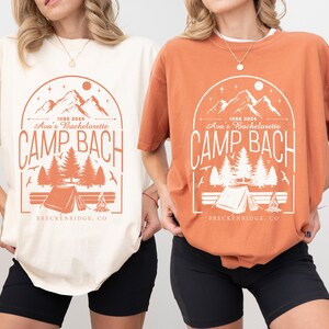 May include: Two t-shirts with a camping theme design. The design features a mountain range, trees, a tent, and a campfire. The text on the shirts reads "JUNE 2024 Ava's Bachelorette CAMP BACH BRECKENRIDGE, CO". One shirt is ivory and the other is orange.
