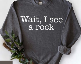 Puede incluir: Una sudadera gris oscuro con el texto "Wait, I see a rock" impreso en blanco en la parte delantera.