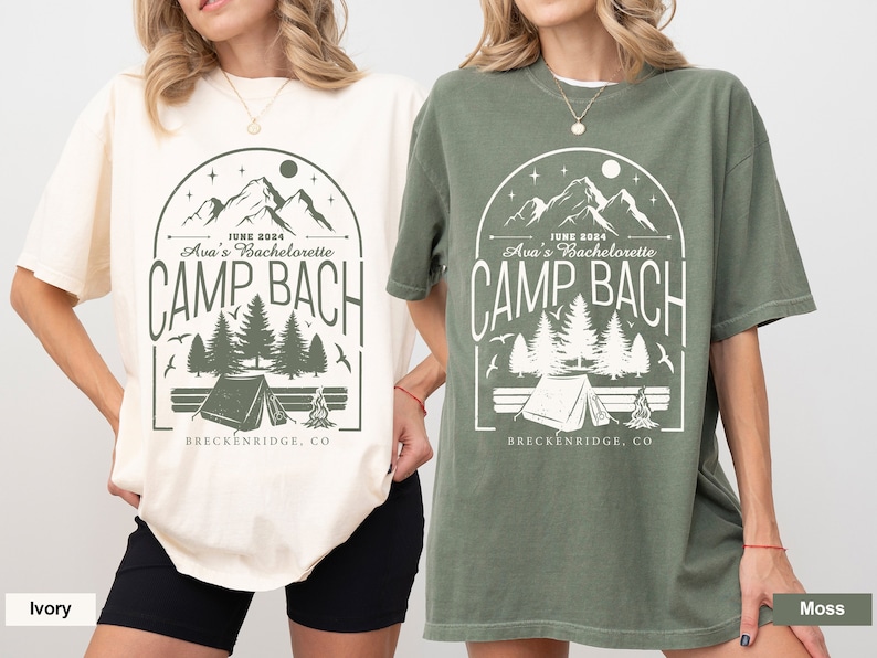 May include: Two t-shirts with a camping theme design. The design features a mountain range, trees, a tent, and a campfire. The text on the shirts reads "JUNE 2024 Ava's Bachelorette CAMP BACH BRECKENRIDGE, CO". The shirts are available in ivory and moss.