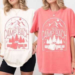 May include: Two t-shirts with a camping theme design. The design features a mountain range, trees, a tent, and a campfire. The text on the shirts reads "JUNE 2024 Ava's Bachelorette CAMP BACH BRECKENRIDGE, CO". The shirts are available in ivory and watermelon.