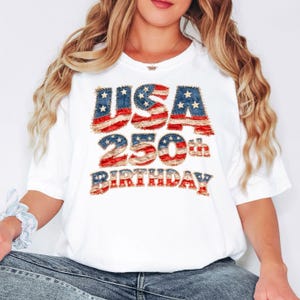 May include: White t-shirt with the text "USA 250th BIRTHDAY" in red, white, and blue, resembling the American flag. The shirt is a crew neck and has short sleeves. The text is in a distressed, vintage style.