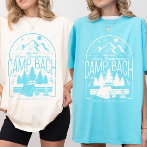 May include: Two oversized t-shirts with a graphic design of a mountain range, trees, a tent, and a campfire. The text on the shirts reads "June 2024 Ava's Bachelorette Camp Bach Breckenridge, CO". The shirt on the left is ivory and the shirt on the right is lagoon blue.