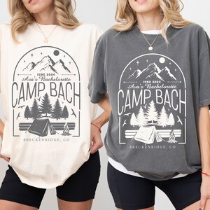May include: Two oversized t-shirts with a camping theme. The shirts are in ivory and pepper colors. The design features a mountain range, trees, a tent, a campfire, and the text "JUNE 2024 Ava's Bachelorette CAMP BACH BRECKENRIDGE, CO".